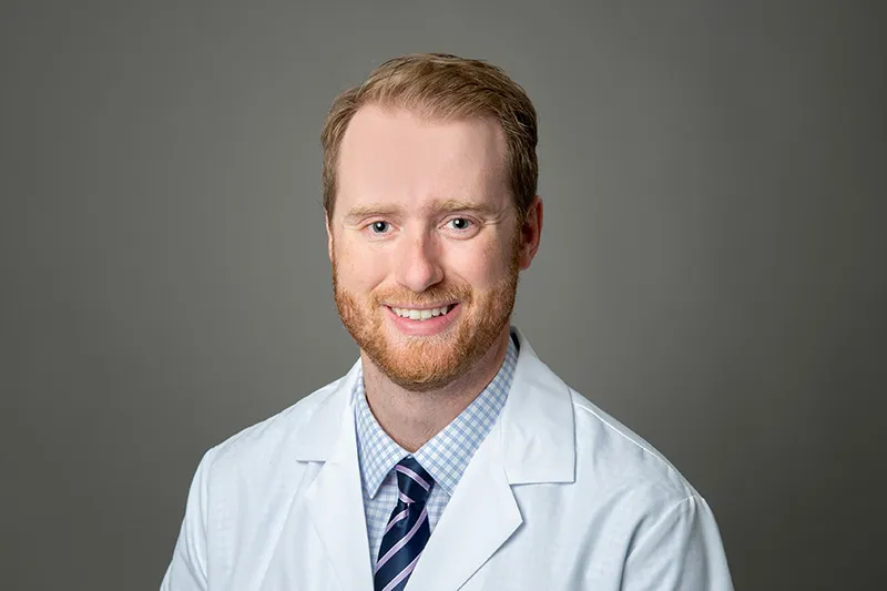 Dr. Whitlock has short strawberry blond hair, beard and mustache, is wearing a light blue dress shirt and tie under a white medical coat and is smiling at the camera