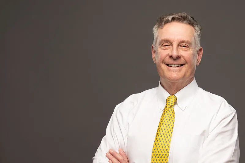 Dr. Pike has short gray hair, is wearing a white dress shirt with yellow tie, and is smiling at the camera.