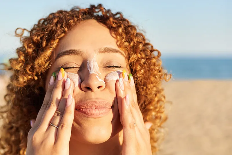 A woman with curly red hair applies sunscreen to her face while at the beach.