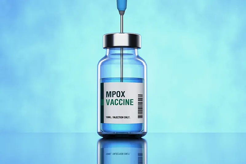 A needle punctures the top of a vial of mpox vaccine against a blue background.