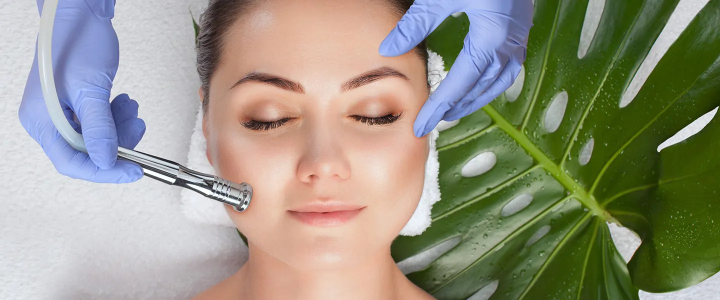 An aesthetician wearing blue surgical gloves performs a facial on a woman with a green palm leaf in the background
