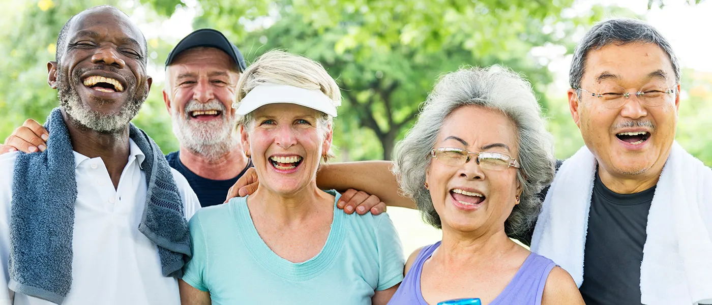 Five older adults of diverse ethnicity smile at the camera. They are in a park surrounded by green grass and trees, and are dressed for fitness activities.