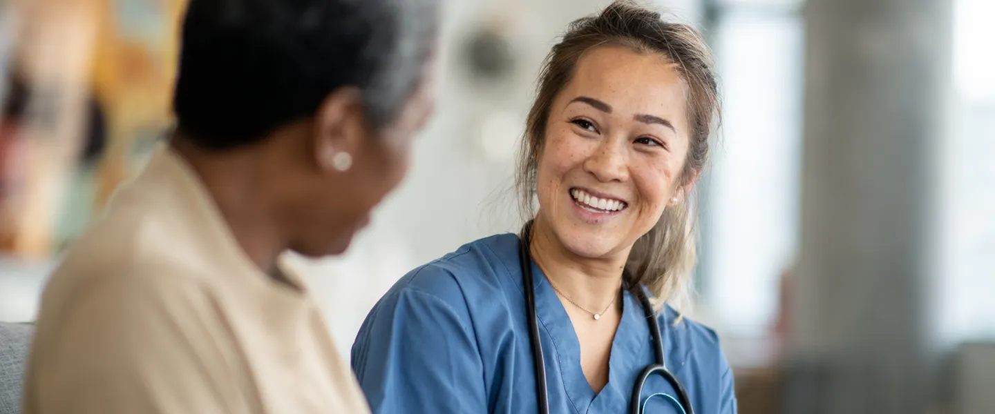 A female nurse sits with a senior patient with stethoscope over her shoulders smiling.