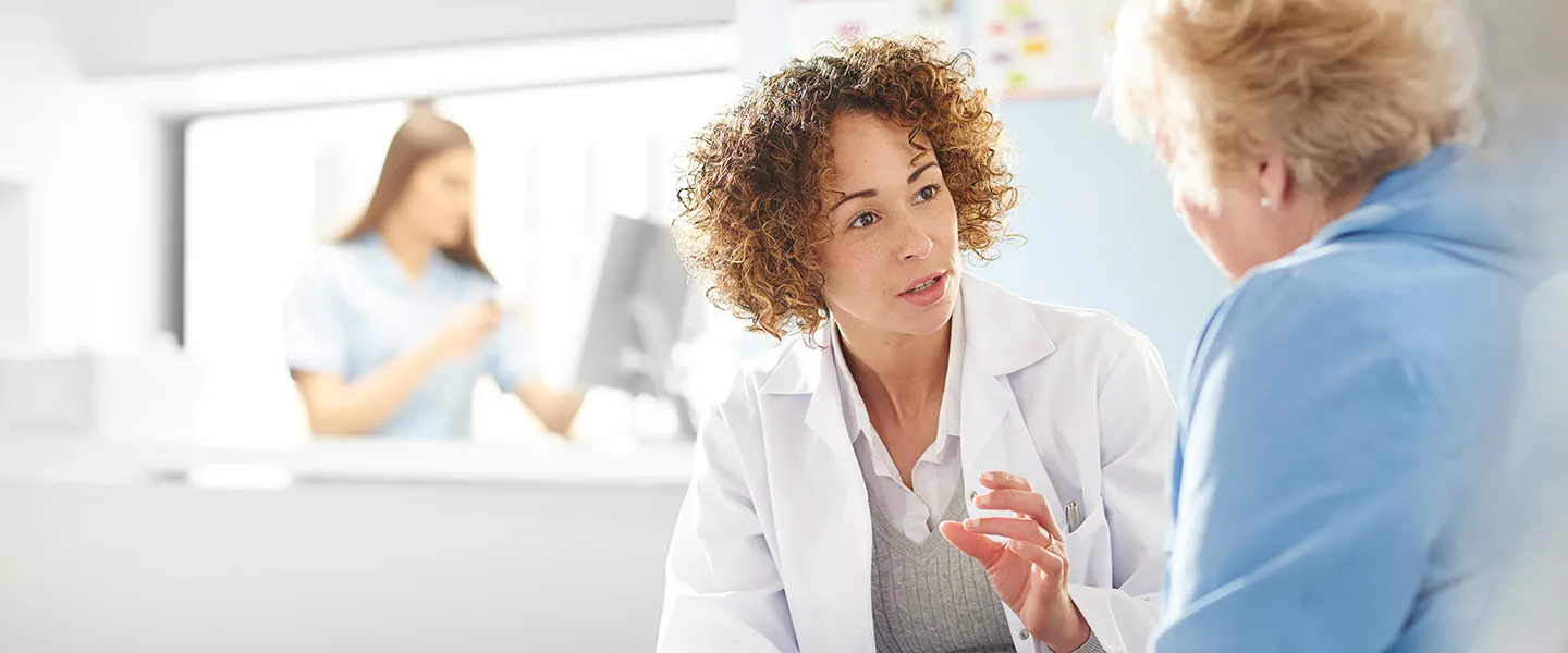 A psychiatrist with short curly hair wears a white medical coat and speaks with an older patient