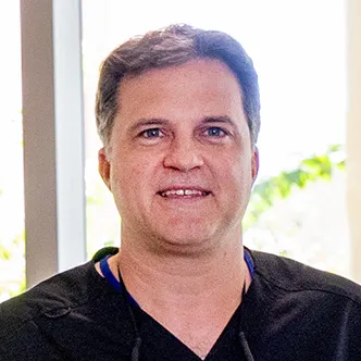 Dr. Bak has short dark hair, is wearing black surgical scrubs, and is smiling at the camera