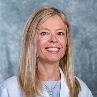 Dr. Margaret Baumgarten is a woman with long blonde hair, wears a white doctor coat and smiles at the camera