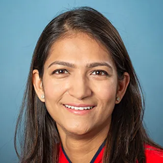 Dr. Patel has long, straight dark hair, is wearing a bright red blouse with a bow around the neck, and is smiling at the camera.