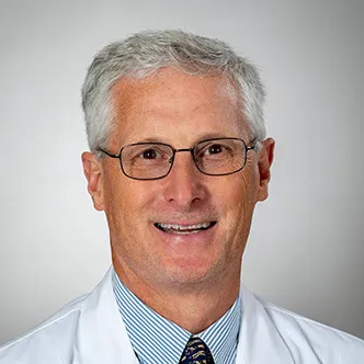 Dr. Sayles has short gray hair, is wearing glasses and a blue striped shirt with dark tie under a white medical coat. He is smiling at the camera.