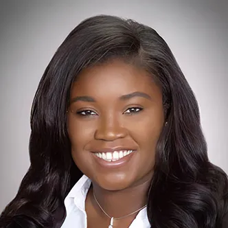 Dr. Cudjoe has long dark hair, is wearing a black jacket and white dress shirt with necklace, and is smiling at the camera