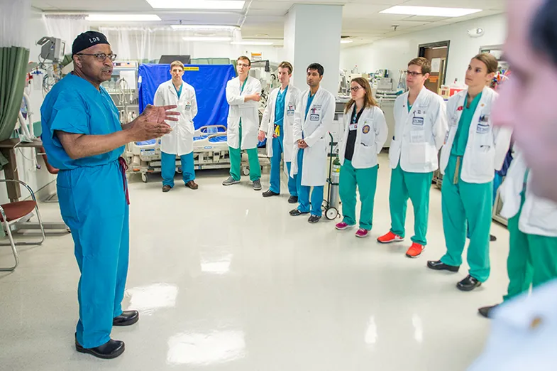 Clad in blue surgical scrubs, Dr Britt stands in a hospital hallway talking to large group of residents.