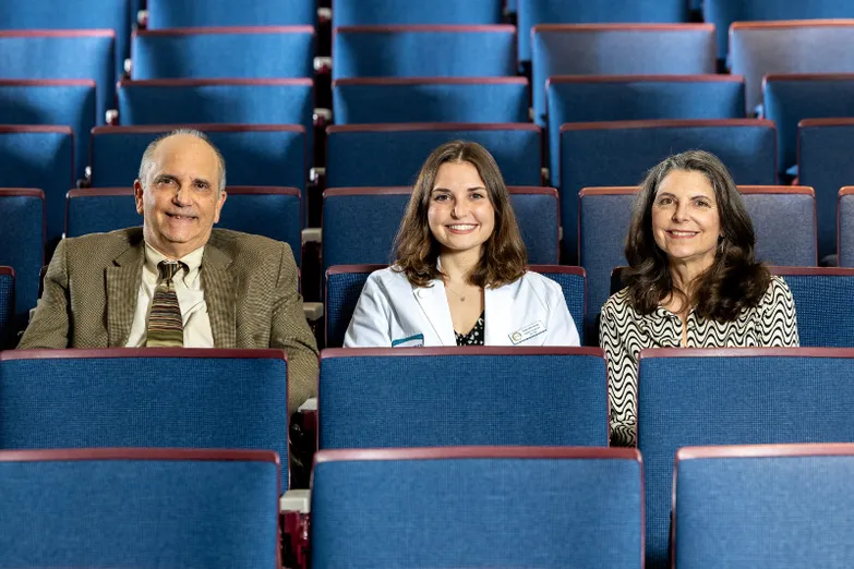 Three individuals smiling while sitting in blue seats in an auditorium.
