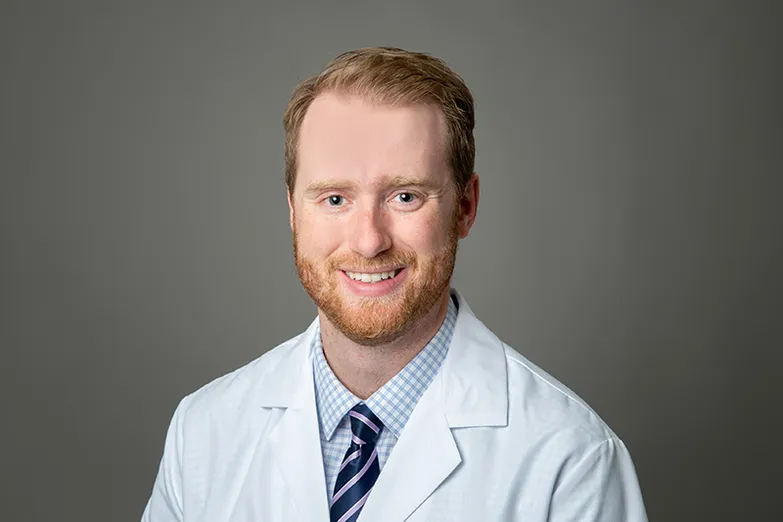 Dr. Whitlock has short strawberry blond hair, beard and mustache, is wearing a light blue dress shirt and tie under a white medical coat and is smiling at the camera