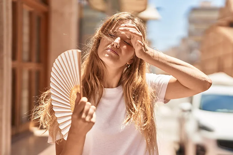 A woman with long blonde hair walks down a street in the summer. She is overheated and is using a handheld fan.
