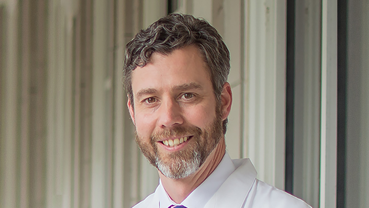 Dr. Dobratz has short brown curly hair and graying goatee, wears a white medical coat, and is smiling at the camera