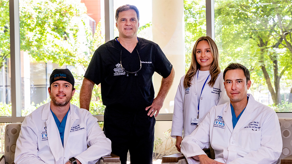 Four surgeons pose in a building atrium with windows behind them. Three wear white medical coats and one is wearing black surgical scrubs.
